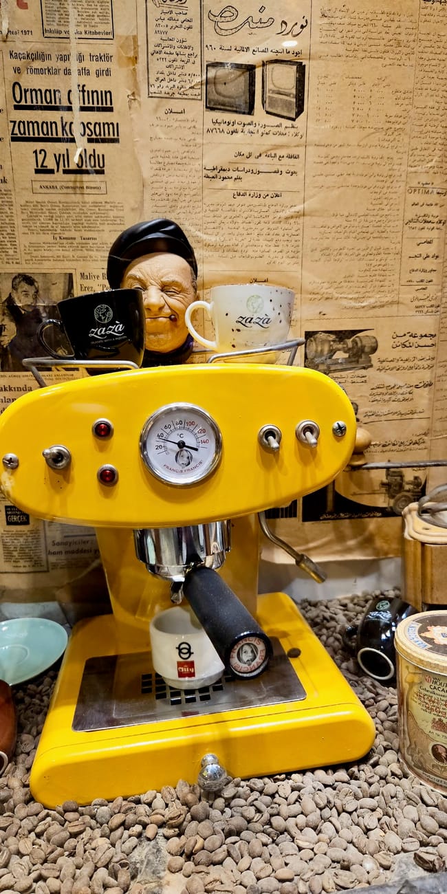Żółty automat ekspresowy z manometrem i kubkami kawy, w tle gazety, płyty i metalowa puszka w stylu retro