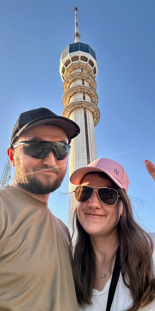 Para w okularach i czapkach selfie na tle wysokiej wieży nad miastem, w słoneczny błękitny dzień