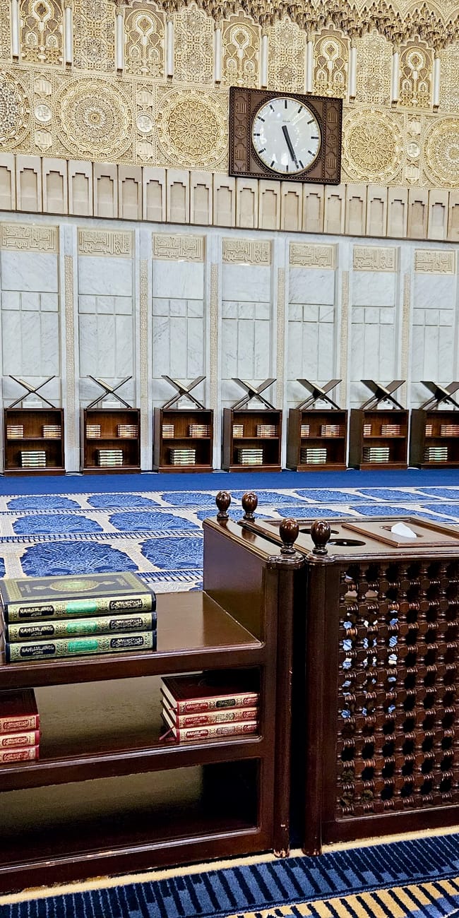 Kuwait city grand mosque prayer hall with ornate clock, arched wall patterns, and blue carpet rows with desks