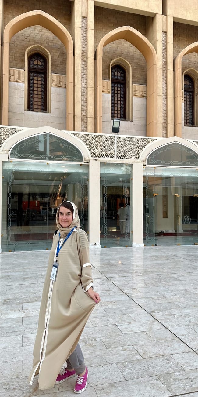 A woman stands in front of kuwait city grand mosque with arched stone entrances and decorative glass panels.