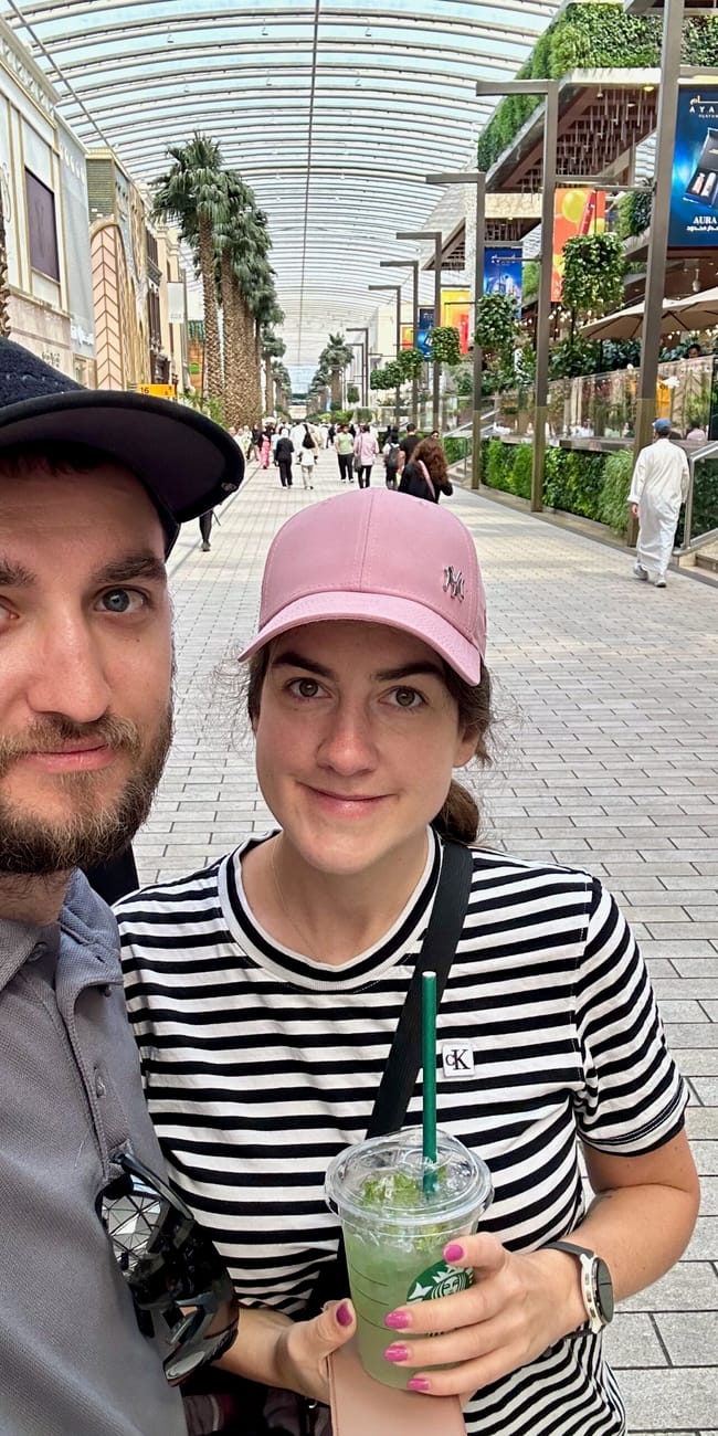 Couple selfie in kuwait city’s the avenues shopping mall, holding a green drink under a glass arcade roof