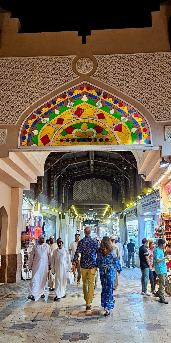 Wejście do muttrah souq w muskacie, z kolorowym łukiem, sklepami po bokach i spacerującymi ludźmi.