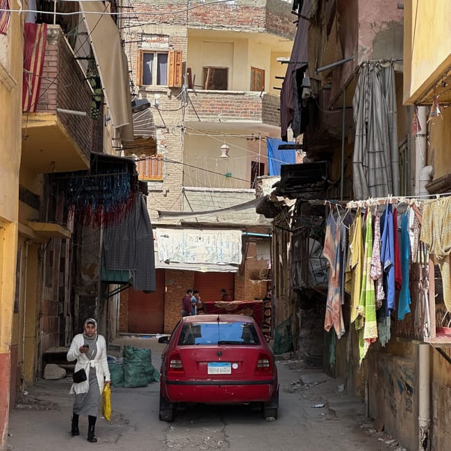 Narrow alley in egypt with a red car, hanging laundry, and a woman walking past.