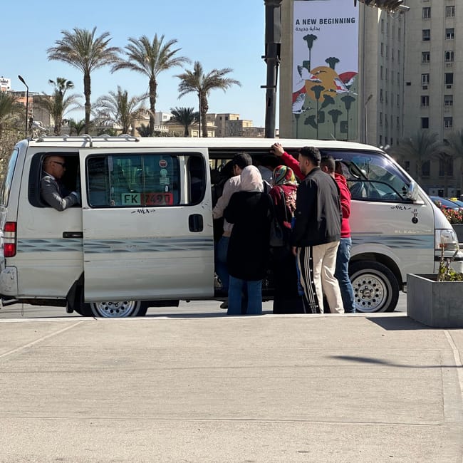 People boarding a white minibus on a street in egypt, with palm trees and buildings in the background