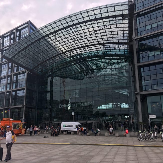 Berlin hauptbahnhof z przeszklonym wejściem i łukowym dachem, przed dworcem stoją piesi i rowery.