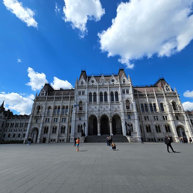 Budapeszteński parlament na tle błękitnego nieba z chmurami i kilkoma spacerowiczami na placu