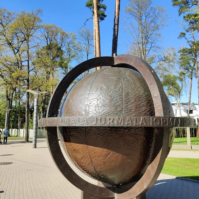 Rzeźba kuli ziemskiej w jurmale, otoczona metalowym pierścieniem na tle drzew i błękitnego nieba