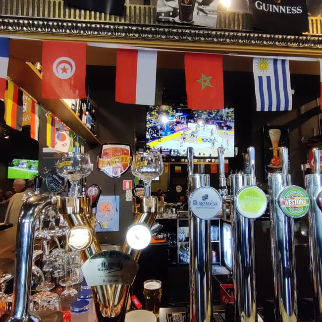 Bar w rydze z beczkami piwa, wiszącymi flagami krajów i telewizorem z meczem.