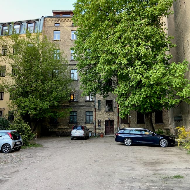 Kamienica w rydze z podwórzem, drzewami i zaparkowanymi samochodami.