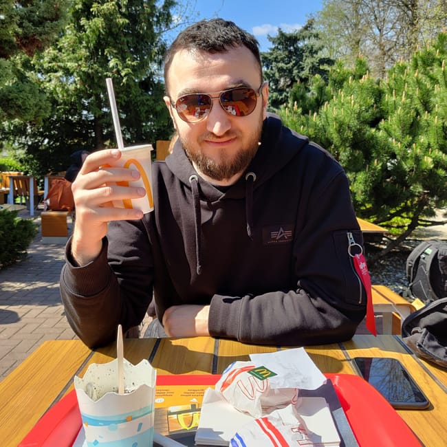 Mężczyzna w okularach przeciwsłonecznych siedzi przy stole na zewnątrz w wilnie, trzymając napój i posiłek z mcdonald’s.