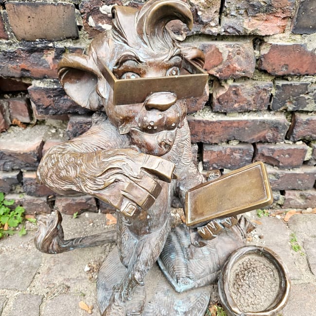 Rzeźba krasnala w gdańsku z tabliczką i ceglaną ścianą w tle