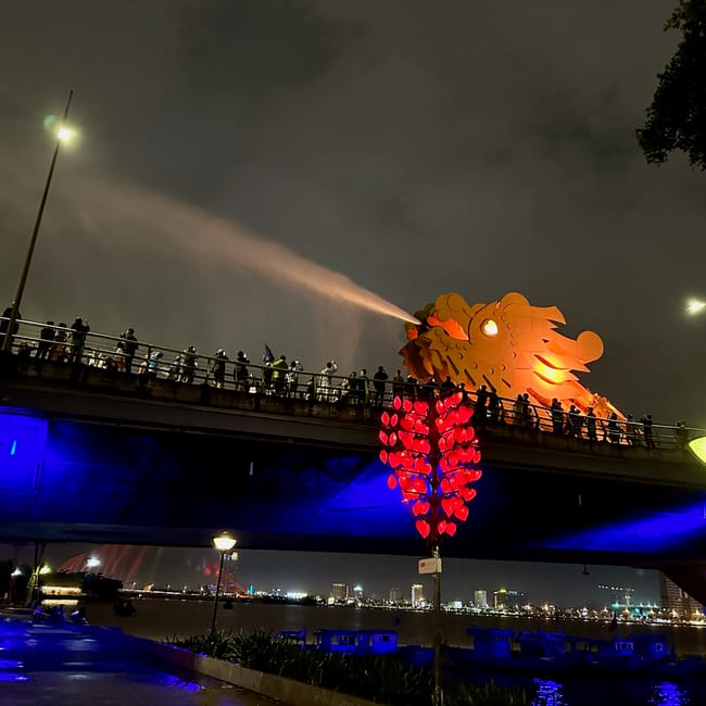 Smoczy most w da nang nocą, z iluminowanym smokiem zionącym wodą i tłumem na moście.