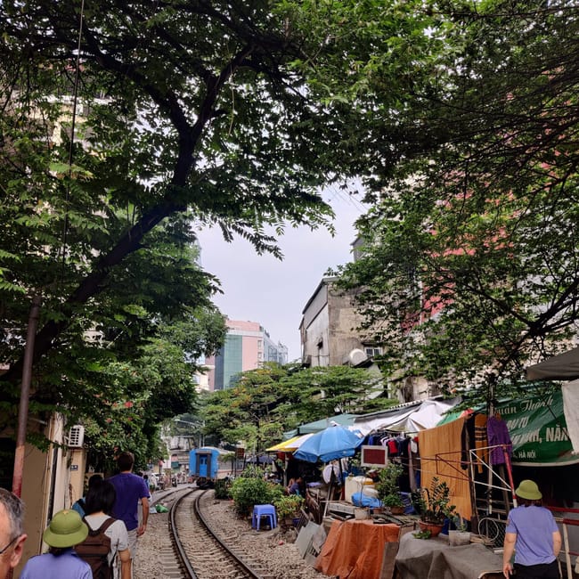 Train street w hanoi, z zakrzywionymi torami, straganami i pieszymi pod gęstymi drzewami.