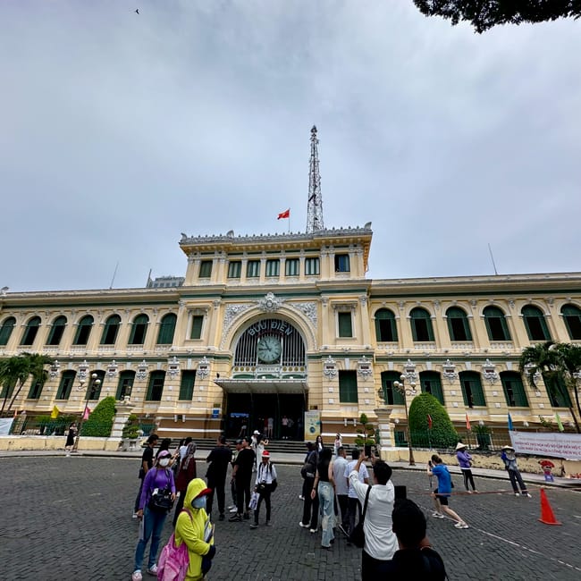 Poczta w ho chi minh z zegarem nad wejściem i turystami na placu.