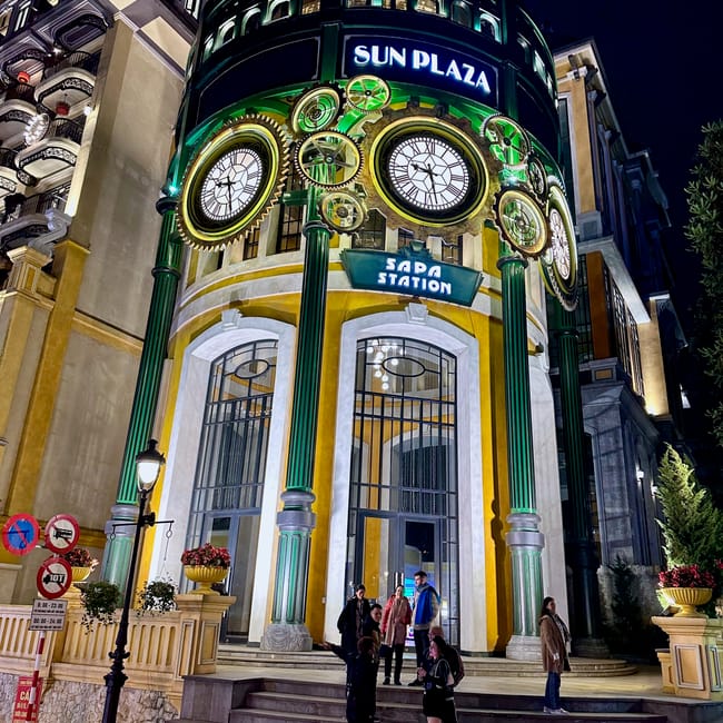 Nocne wejście do sun plaza w sa pa z dużym zegarem i kilkoma osobami przed budynkiem.