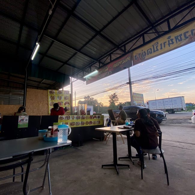 People dining at an open-air street stall in prachinburi, thailand, with cars parked nearby at sunset