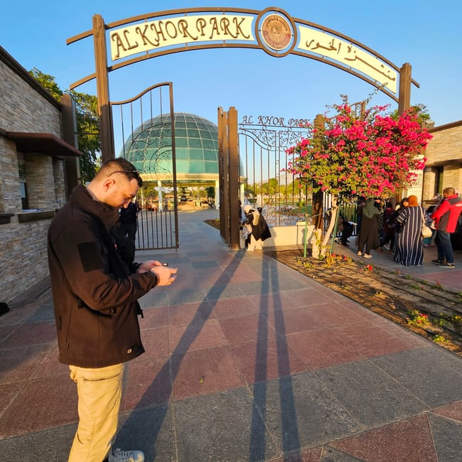 Wejście do al khor park w doha, z odwiedzającymi i kwitnącym krzewem.
