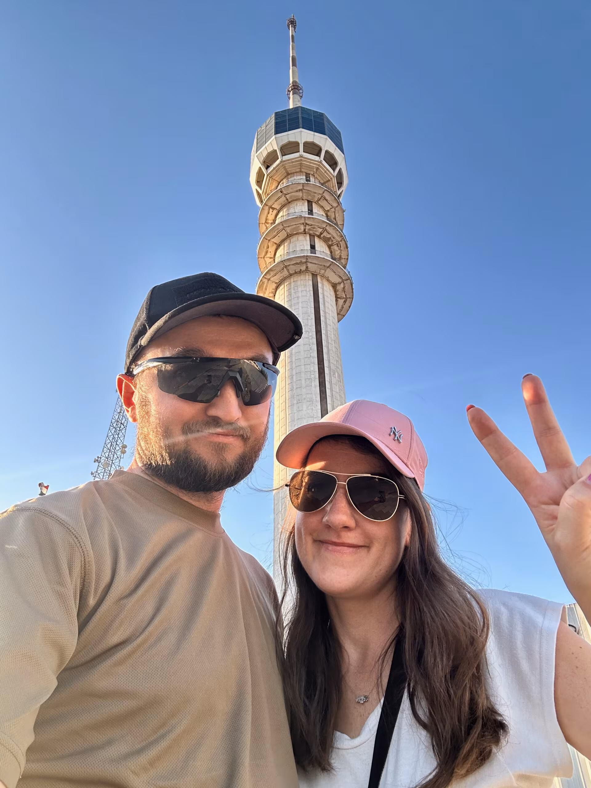 Uśmiechnięta para w okularach i czapkach przed wieżą eiffla; nad nimi błękitne niebo, selfie z uniesioną dłonią