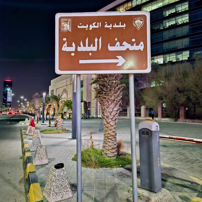 Kuwait city road sign with arabic text, arrow pointing right, in front of a lit building at night