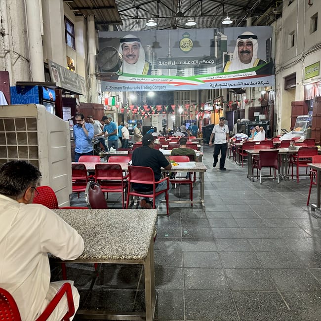 Indoor restaurant in kuwait, city al mubarakiya, with red chairs and a banner featuring two men in traditional dress