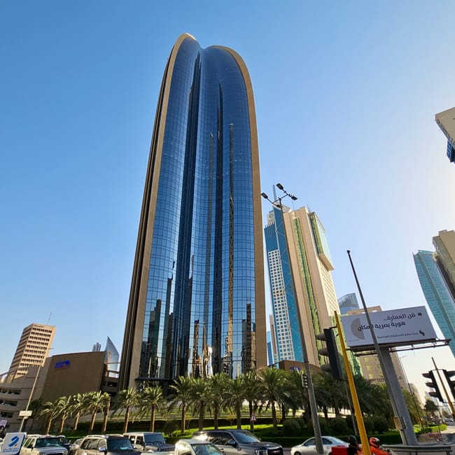 Kuwait city street photo featuring a tall modern glass skyscraper and parked cars under a blue sky