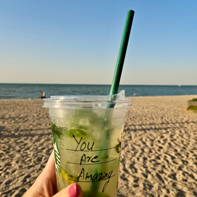 Kuwait city waterfront: hand holding a green iced drink with a green straw on a sandy beach