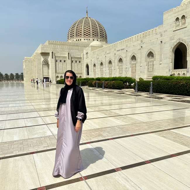 Kobieta na dziedzińcu muscat grand mosque w omanie, na tle jasnego budynku z kopułą i minaretem