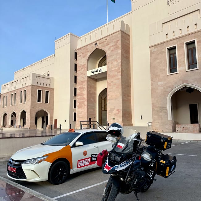 Motocykl podróżny przed budynkiem mof w muskacie w omanie, obok taksówki yango i pod flagą omanu