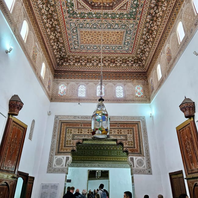 Bahia palace in marrakesz, morocco, showing an ornate moroccan hall ceiling and hanging lantern with visitors