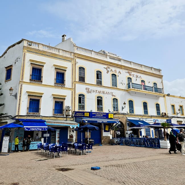Essaouira, plac moulay hassana: biała kamienica z niebieskimi oknami i restauracjami oraz ogródkami z parasolami