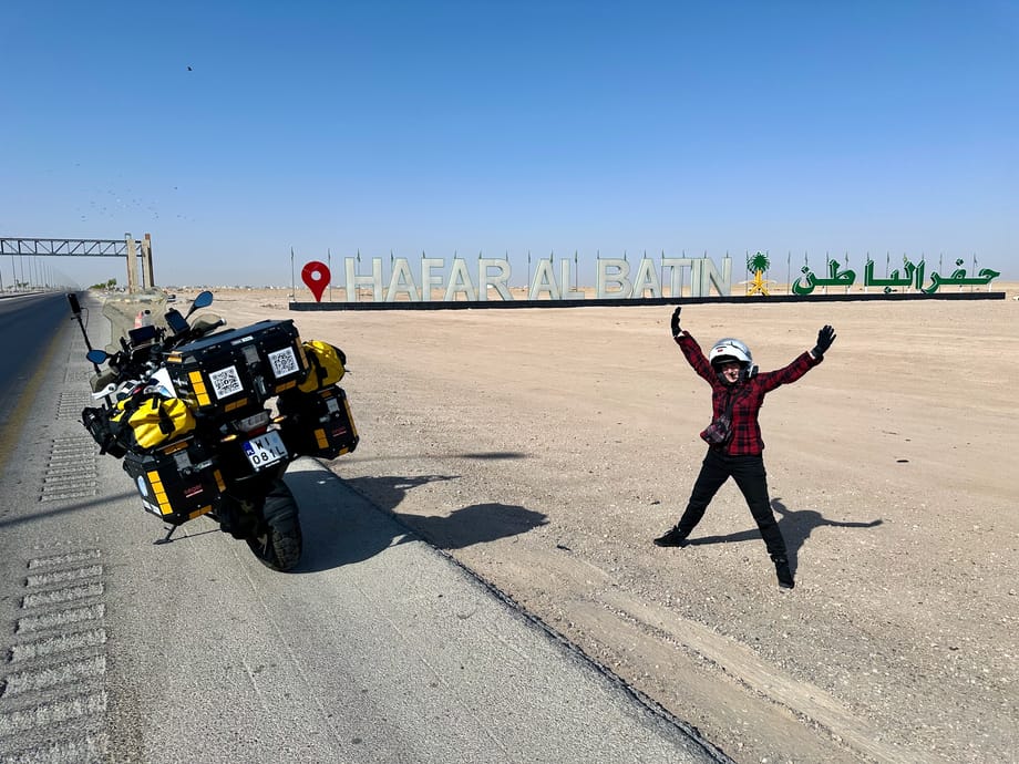 Motocykl wyprawowy przy napisie hafar al batin w arabii saudyjskiej, obok stojący motocyklista.