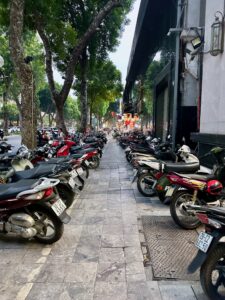 Ulica w hanoi zastawiona zaparkowanymi skuterami przy chodniku i drzewach.