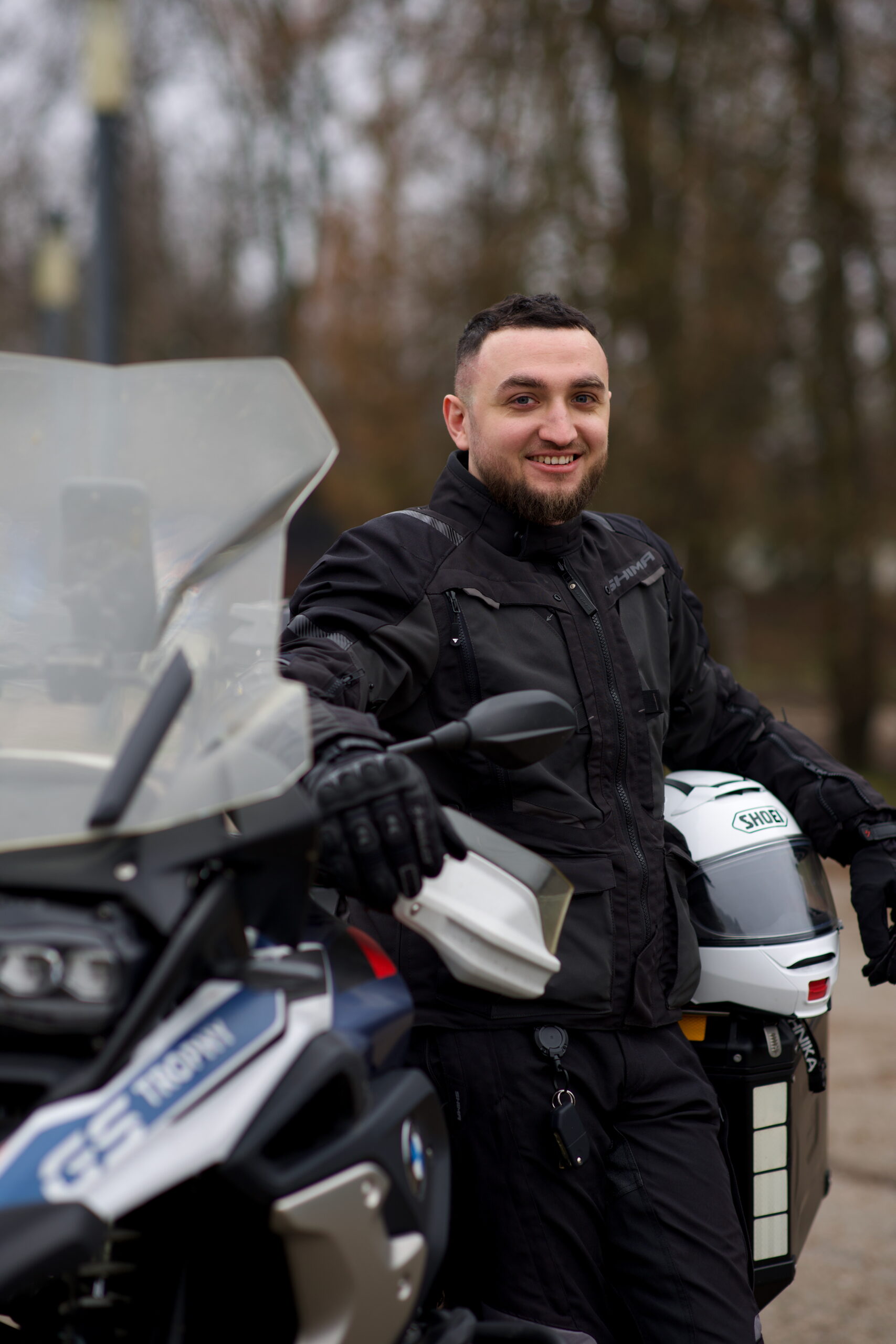 Uśmiechnięty motocyklista w czarnym stroju stoi przy motocyklu turystycznym na tle drzew.