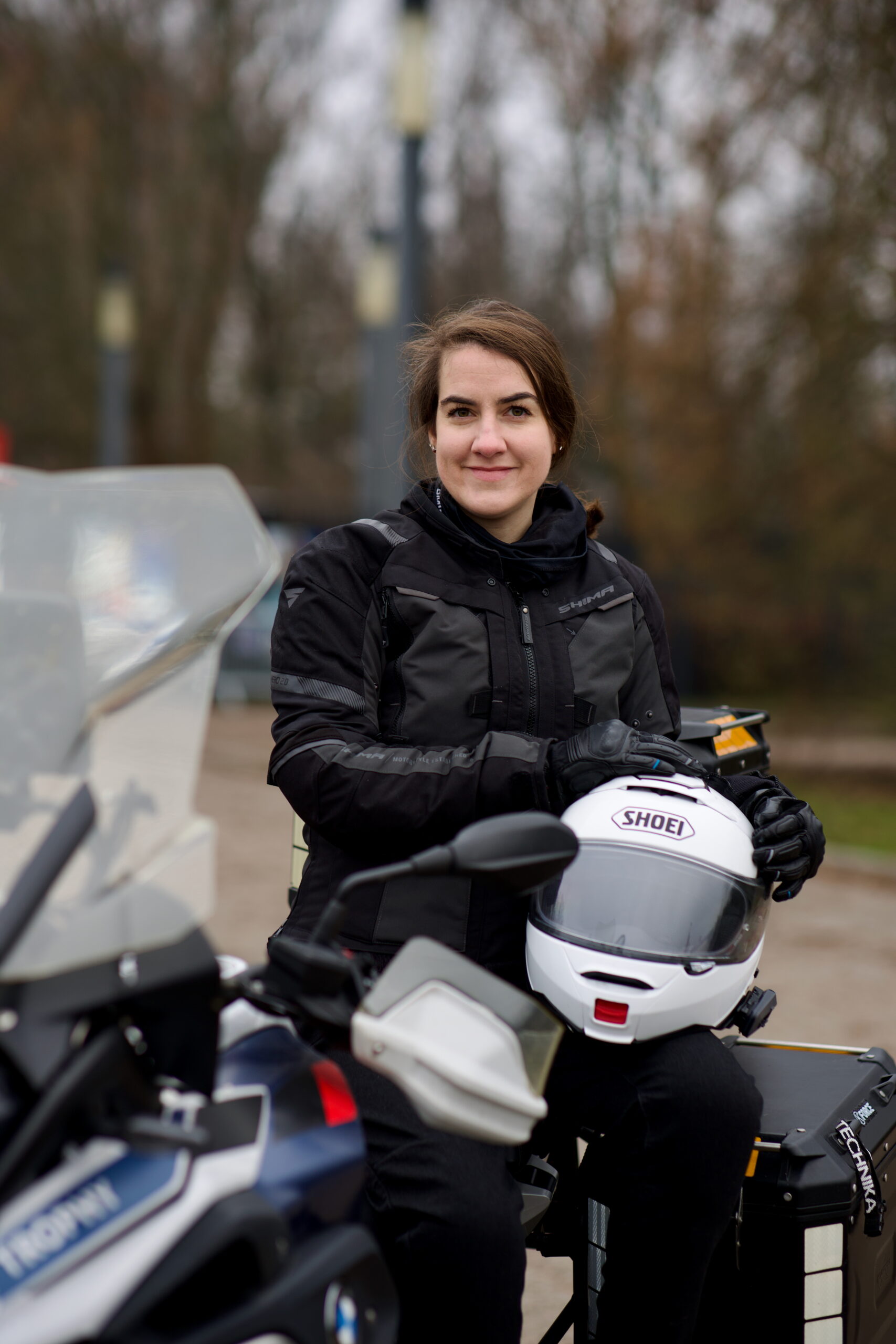 Kobieta w motocyklowym stroju siedzi na motocyklu i trzyma biały kask shoei.