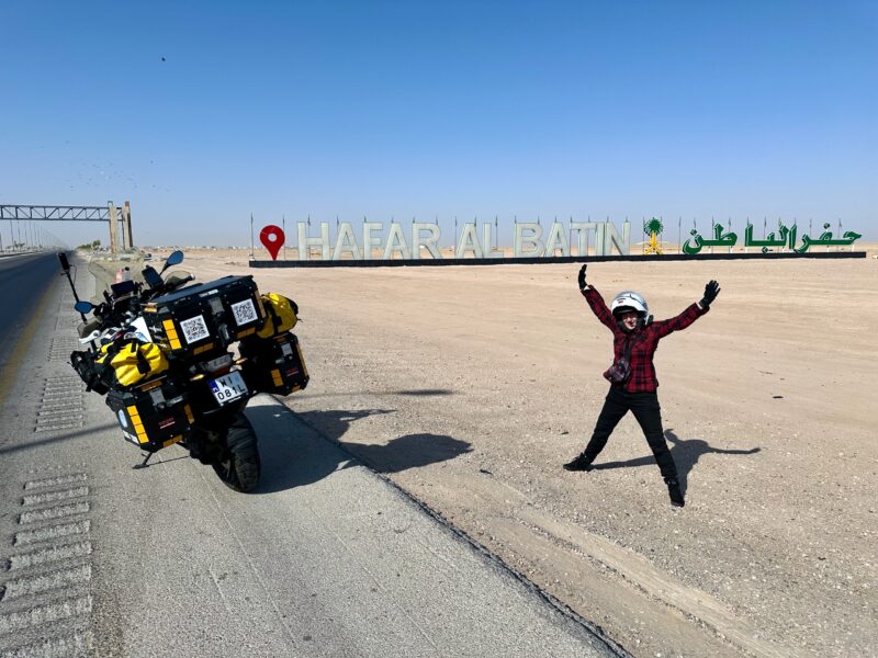 Motocykl wyprawowy przy napisie hafar al batin w arabii saudyjskiej, obok stojący motocyklista.