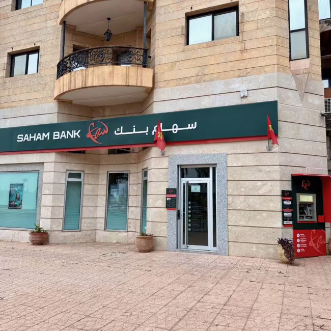 Saham Bank na parterze nowoczesnego kamienistego budynku, z wejściem z lewej strony i bankomatem po prawej, nadzwyczajne okna i balkon nad wejściem.