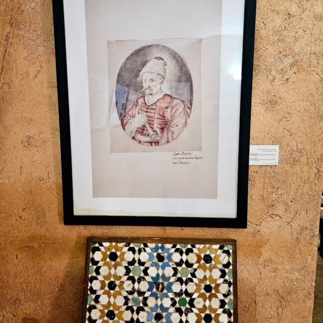 Portret starca w turbanie, z przodu ręką na piersiach, utrwalony w efemerycznym kolażu, na tle ozdobny mozaikowy panel.