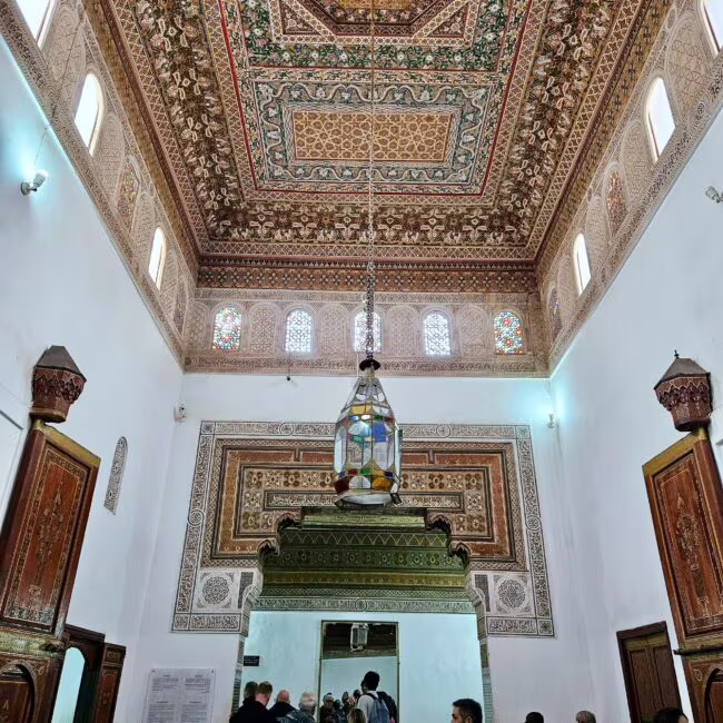 Wnętrze meczetu z misterną arabeską na suficie i mihrabem, nad tłumem ludzi zawieszona kolorowa lampa