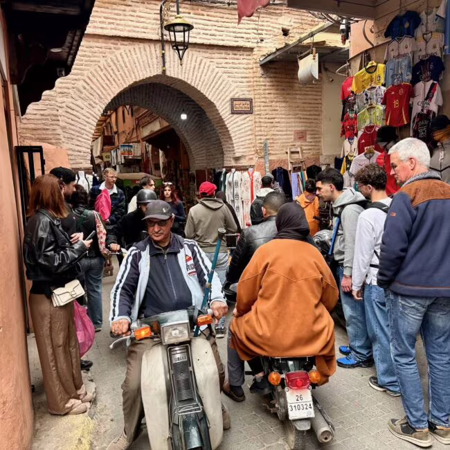 Wąska uliczka handlowa z ceglaną architekturą, tłum ludzi na chodniku, stoją skutery, w prawej części sklep z odzieżą na wieszaku, arch podcieniowy w tle.