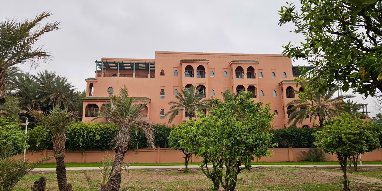 Południowa ściana różowego, wielopiętrowego budynku w oranżowej tonacji, otoczonego palmami i ogrodem z krzewami.