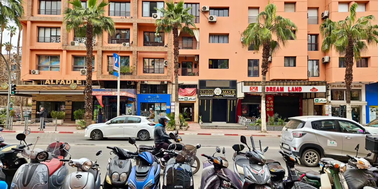 Ulica z różowo-orangistym blokowiskiem, w dole sklepy i tablice, w pierwszym planie rząd skutera i motorów, po środku palmowe drzewa.