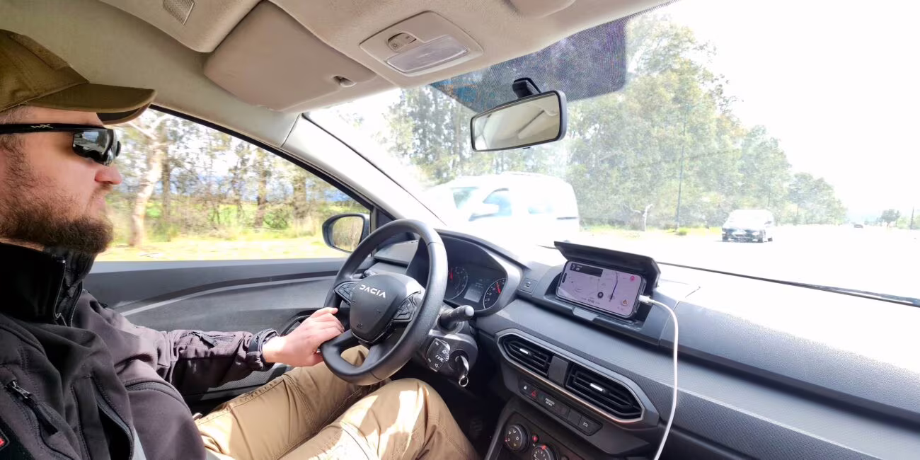 Kierowca z brodą i ochronnymi okularami siedzi za kierownicą w czarniej kurtce, prowadzi samochód na szybkiej drodze, na desce widnieje uchwyt z telefonem.