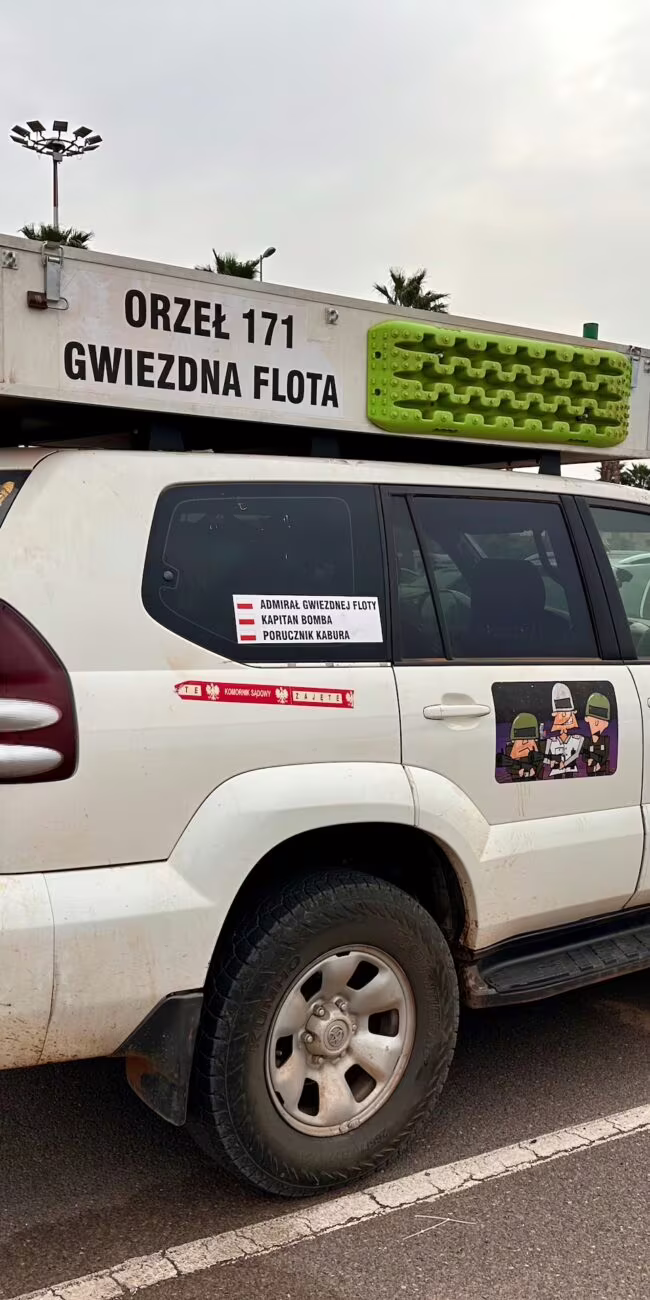 Samochód terenowy z bagażnikiem na dachu i załadowanym sprzętem, w plenerowym parku palmowym, z numerem 171 i reklamami.