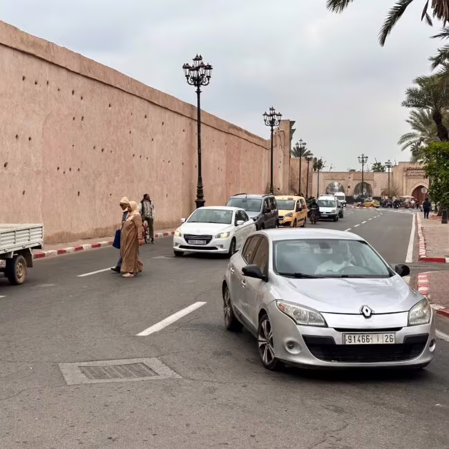 Szara Renault na ruchliwej ulicy przy wysokiej, różowej kamiennej mury miejskiej, ludzie przechodzą przed nią, po lewej stoi biała ciężarówka, po prawej palmowe drzewo i chodnik.