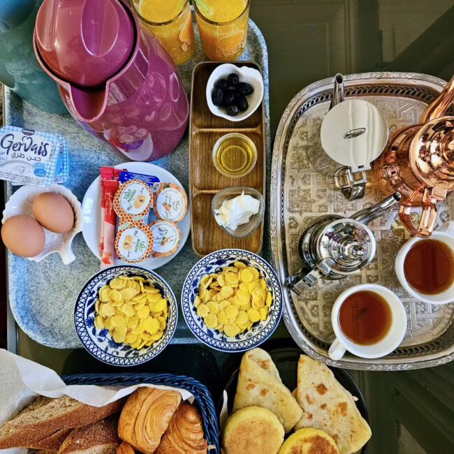 Śniadaniowy stół z rogalikami, bułkami i placuszkami, miseczkami płatków, jajkami, dżemem, serkami i sokami, obok czajnik i kubki z herbatą.