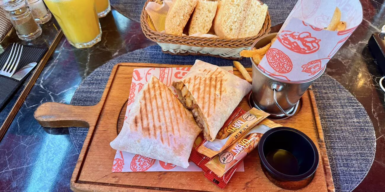 Pierś z grillowaną tortillą, chipsy w metalowym kubku, sosy i różne przyprawy na drewnianej desce; obok kosz z chlebem i dwa soki pomarańczowe.