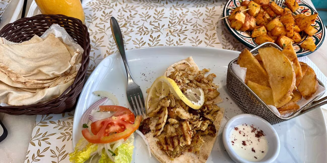 Tortilla z grillowaną mieszanką mięsno-warzywną, plasterkiem cytryny, cebulą, pomidorem i sałatą na talerzu, obok frytki z sosie i miska sosu, w oknie zapiekanki.