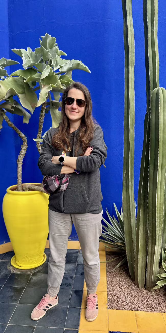 Osoba stojąca na tle intensywnie niebieskiej ściany, ubrana w szarą bluzę, okulary przeciwsłoneczne i jasne spodnie, z różowymi butami, obok żółta doniczka z dużym roślinnym krzewem.