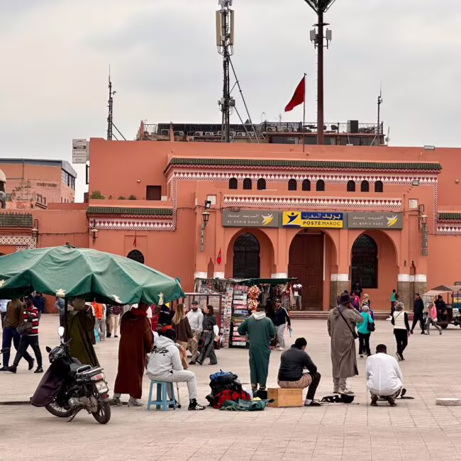 Piazza przed fasadowym, ceglastopomarańczowym budynkiem bankowym z arkadowymi wejściami, gdzie ludzie odpoczywają, stoją pod zielonym parasolem, a miejski tłum spaceruje.
