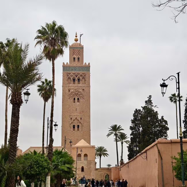 Wieża wznosząca się nad promenadą z palmami i ludźmi w tunelowej bramie w Marrakeszu